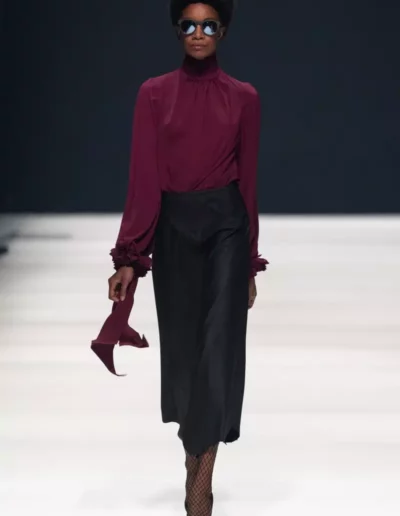 Ein Model läuft in einem burgunderfarbenen Oberteil und einem schwarzen Rock über den Laufsteg.