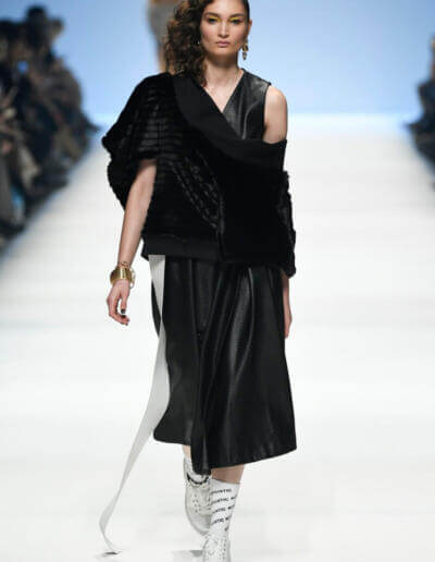 Ein Model läuft in einem schwarzen Kleid und einem Pelzmantel über den Laufsteg.