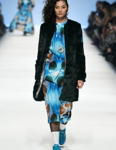 Ein Model läuft in einem blauen Mantel und einem Kleid mit Blumendruck über den Laufsteg.
